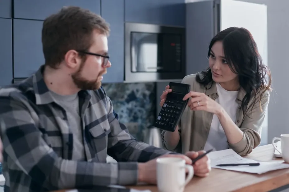 A couple discussing finances with a calculator and papers at a wooden table, looking concerned.